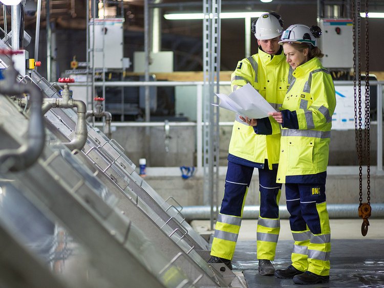 Photo: one female and one male employee, wearing work clothes and concentrating on printed documents; at left, the raking system of the existing sewage treatment plant.