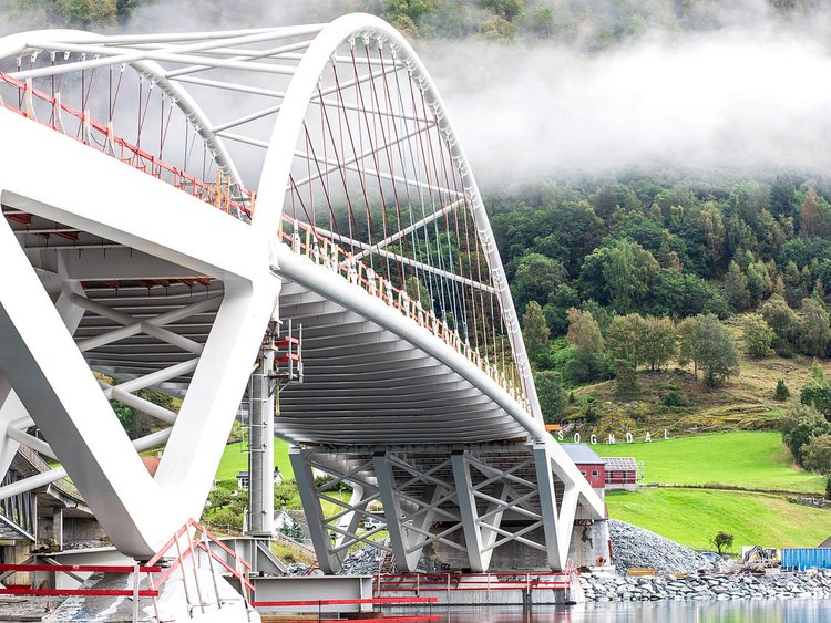 Photo: View of the bridge from below with the two pile heads in the fjord; in the right of the image is an embankment with meadows, a fog-shrouded forest and a few houses.