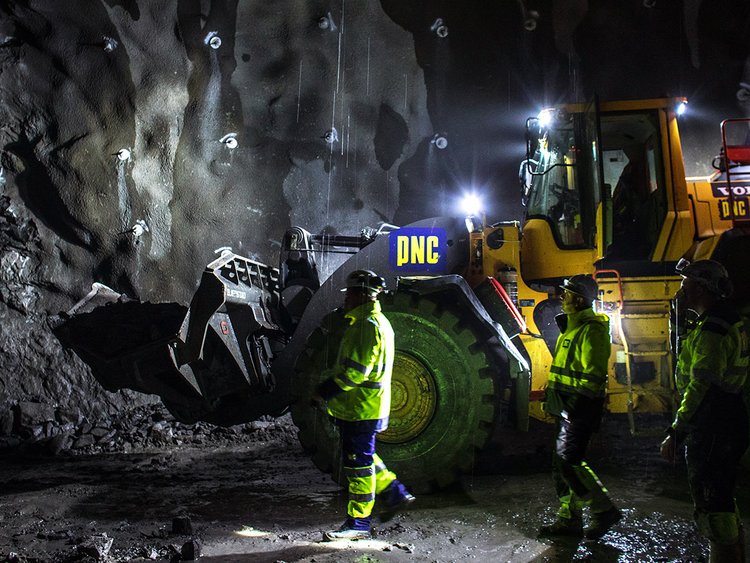 Photo: three tunneling workers in working clothes and helmet walking towards a dark rock face, an excavator and a tunnel wall sprayed with concrete in the background