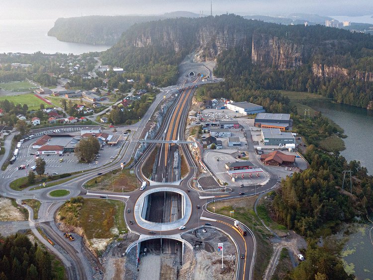 Aerial photo: traffic junction consisting of newly built bridges, roundabouts and a tunnel on the outskirts of a city, water surfaces and mountains on the right and in the back.